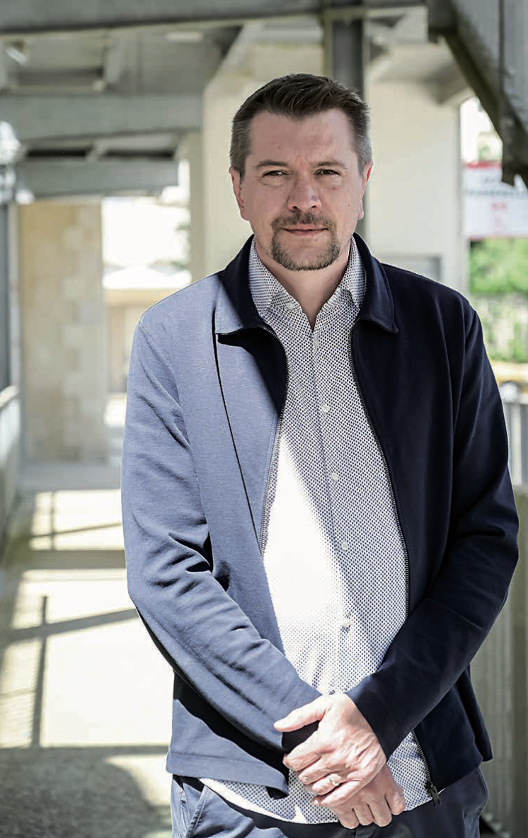 Ruben Boisseleau, un homme aux cheveux bruns courts et à la barbe taillée, se tient à l'extérieur, sous une passerelle couverte, vêtu d'une veste marine sur une chemise à motifs. Il regarde la caméra avec une expression neutre et les mains jointes devant lui. - Édition Offset 5 (Partenaire de votre communication locale en Vendée)