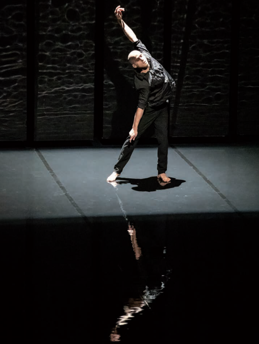Sur une scène faiblement éclairée, un danseur pieds nus, vêtu de noir, se produit dans le style expressif de Ruben Boisseleau, un bras tendu vers le haut et l'autre vers le bas, projetant un reflet dramatique sur le sol brillant. - Édition Offset 5 (Partenaire de votre communication locale en Vendée)