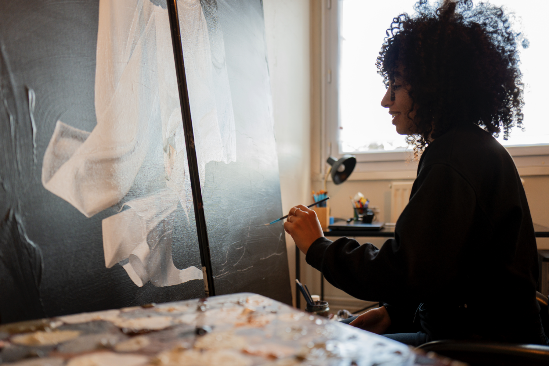 Une personne aux cheveux bouclés est assise à l'intérieur, peignant une grande œuvre d'art abstraite en noir et blanc sur une toile, rappelant le style expressif de Simon Boisliveau, avec du matériel d'art et une fenêtre visible à l'arrière-plan. - Édition Offset 5 (Partenaire de votre communication locale en Vendée)