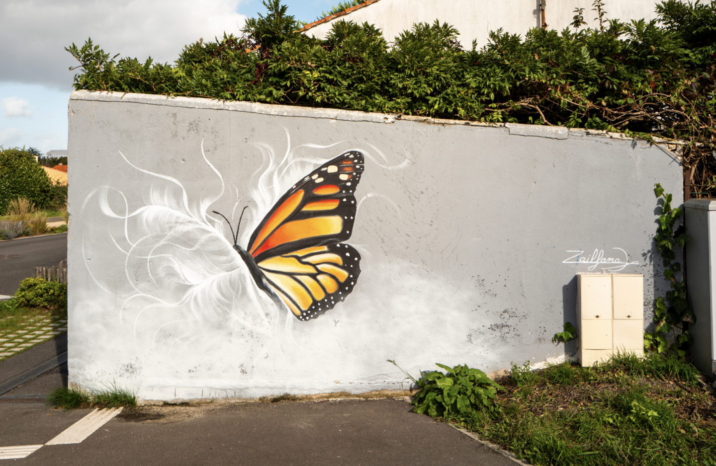 Une peinture murale de Simon Boisliveau représente un papillon monarque aux ailes orange vif, noires et blanches peintes sur un mur extérieur gris. Des lignes blanches ondulées créent un mouvement autour du papillon, tandis que des plantes vertes poussent à la base du mur. - Édition Offset 5 (Partenaire de votre communication locale en Vendée)
