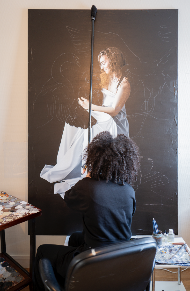 Une personne aux cheveux bouclés est assise devant un chevalet et peint sur une grande toile une image réaliste d'une femme tenant un cygne, inspirée par Simon Boisliveau. Des fournitures artistiques sont disposées sur une petite table à proximité. - Édition Offset 5 (Partenaire de votre communication locale en Vendée)