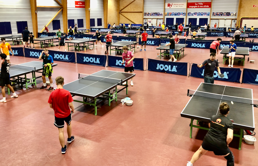 Un grand gymnase intérieur avec plusieurs parties de tennis de table en cours. Des personnes de tous âges, souvent en tenue de sport, jouent ou regardent autour de tables noires séparées par des barrières bleues à Grimpe Atlantique Ouest. - Édition Offset 5 (Partenaire de votre communication locale en Vendée)