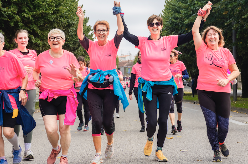 Un groupe de femmes portant des chemises roses assorties sourient et lèvent les bras en signe de célébration alors qu'elles participent à la course à pied en plein air Grimpe Atlantique Ouest dans une rue bordée d'arbres. - Édition Offset 5 (Partenaire de votre communication locale en Vendée)