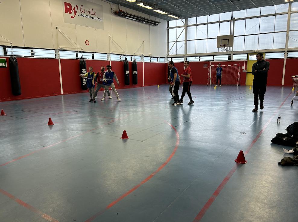 Un groupe de personnes pratique un sport en salle dans un gymnase aux murs rouges et aux grandes fenêtres à Grimpe Atlantique Ouest. Plusieurs cônes délimitent le sol, tandis que des sacs de boxe sont accrochés au mur et qu'un homme regarde près du but. - Édition Offset 5 (Partenaire de votre communication locale en Vendée)