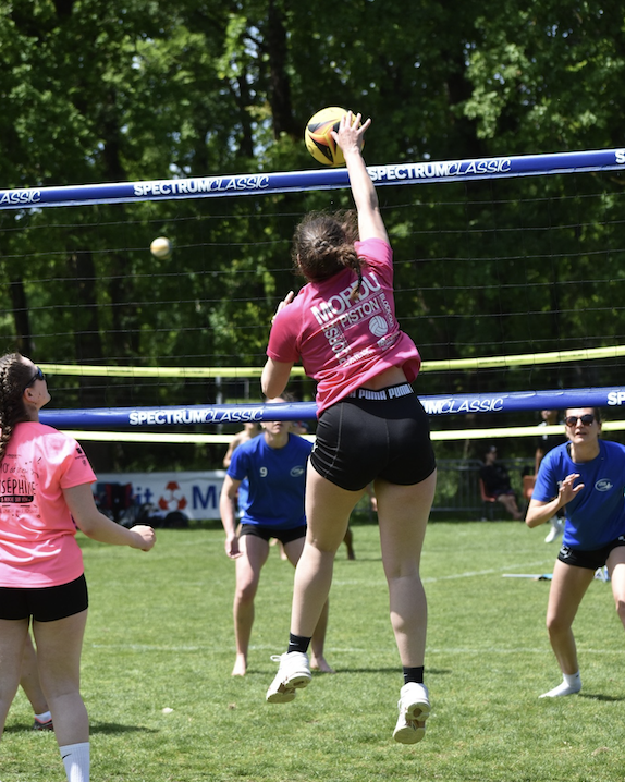 Un joueur de volley-ball en chemise rose saute pour frapper le ballon au-dessus du filet lors d'un match en plein air de Grimpe Atlantique Ouest, tandis que ses coéquipiers et adversaires en chemise bleue et rose regardent et se préparent sur le terrain herbeux. - Édition Offset 5 (Partenaire de votre communication locale en Vendée)