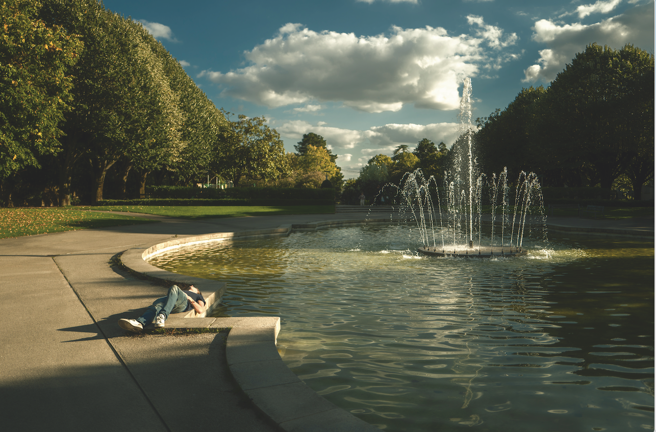Une personne est allongée sur un banc de pierre incurvé à côté d'une grande fontaine de Veillon dans un parc, entouré d'arbres, sous un ciel partiellement nuageux, la lumière du soleil projetant des ombres chaudes. - Édition Offset 5 (Partenaire de votre communication locale en Vendée)