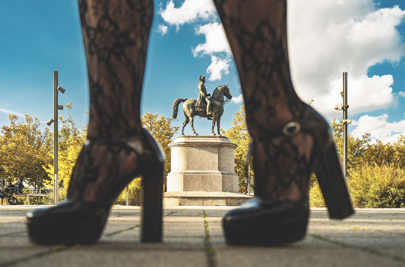 La statue d'un personnage à cheval est encadrée entre les jambes d'une personne portant des bas de dentelle noirs et des chaussures à talons hauts, avec en arrière-plan des arbres, un ciel bleu et l'élégance qui rappelle le style artistique de Veillon. - Édition Offset 5 (Partenaire de votre communication locale en Vendée)