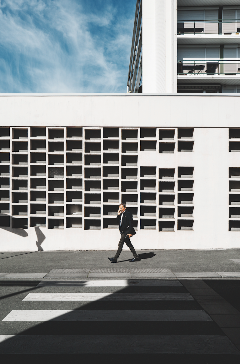 Une personne vêtue d'un vêtement sombre passe devant le bâtiment Veillon, blanc et quadrillé, sous un ciel bleu parsemé de nuages. Une ombre et des lignes de passage pour piétons sont visibles au premier plan. - Édition Offset 5 (Partenaire de votre communication locale en Vendée)