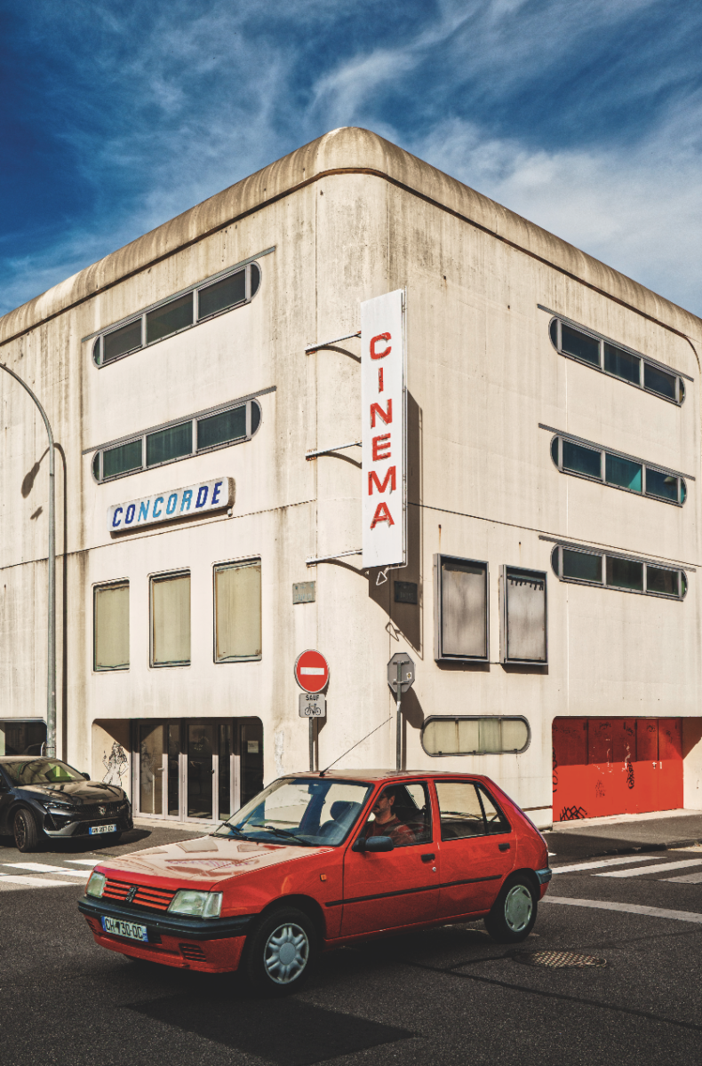 Une voiture rouge passe devant le bâtiment blanc et moderniste du cinéma Veillon, avec une enseigne verticale CINEMA et CONCORDE au-dessus de l'entrée. Le ciel est partiellement nuageux et une autre voiture noire est garée à proximité. - Édition Offset 5 (Partenaire de votre communication locale en Vendée)