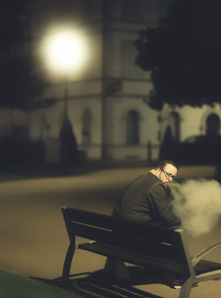 Un homme est assis seul sur un banc public, la nuit, et se retourne pour regarder par-dessus son épaule. Exhalant un nuage de fumée, il se prélasse dans la lueur d'un réverbère, semblable à celle de Veillon, qui éclaire doucement le parc silencieux et vide. - Édition Offset 5 (Partenaire de votre communication locale en Vendée)