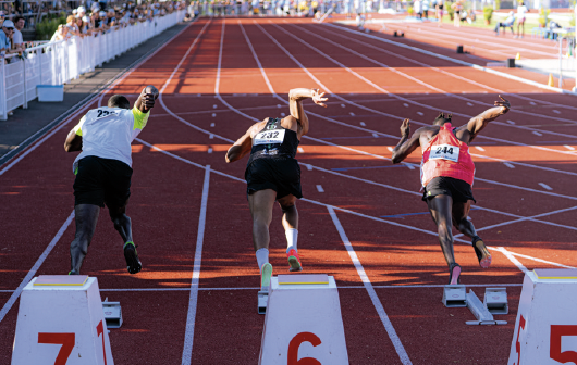 Trois sprinters masculins en tenue de sport s'élancent des blocs de départ sur une piste rouge à La Roche-sur-Yon ville active et sportive, s'élançant dans les couloirs 5, 6 et 7 sous le regard enthousiaste des spectateurs depuis les tribunes. - Édition Offset 5 (Partenaire de votre communication locale en Vendée)