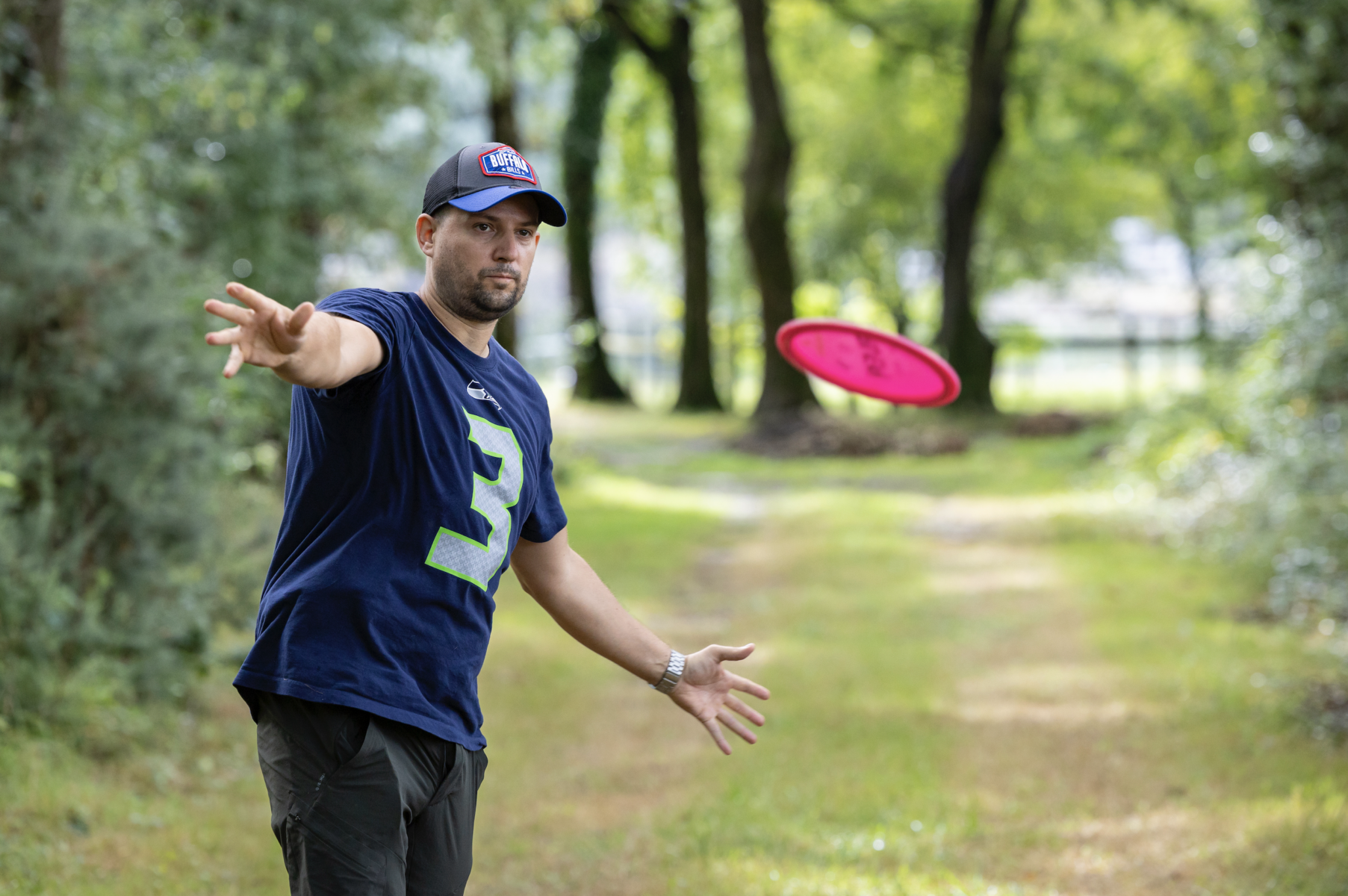 Un homme portant une casquette bleue et un t-shirt bleu marine avec un grand numéro 3 vert lance un frisbee rose dans un parc boisé par une journée ensoleillée, capturant l'esprit de La Roche-sur-Yon ville active et sportive. - Édition Offset 5 (Partenaire de votre communication locale en Vendée)