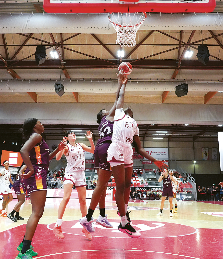 Deux joueuses de basket-ball sautent pour atteindre le ballon près du panier, tandis que leurs coéquipières les regardent attentivement sur un terrain couvert très éclairé à La Roche-sur-Yon ville active et sportive. Des supporters et des bannières sont visibles à l'arrière-plan. - Édition Offset 5 (Partenaire de votre communication locale en Vendée)