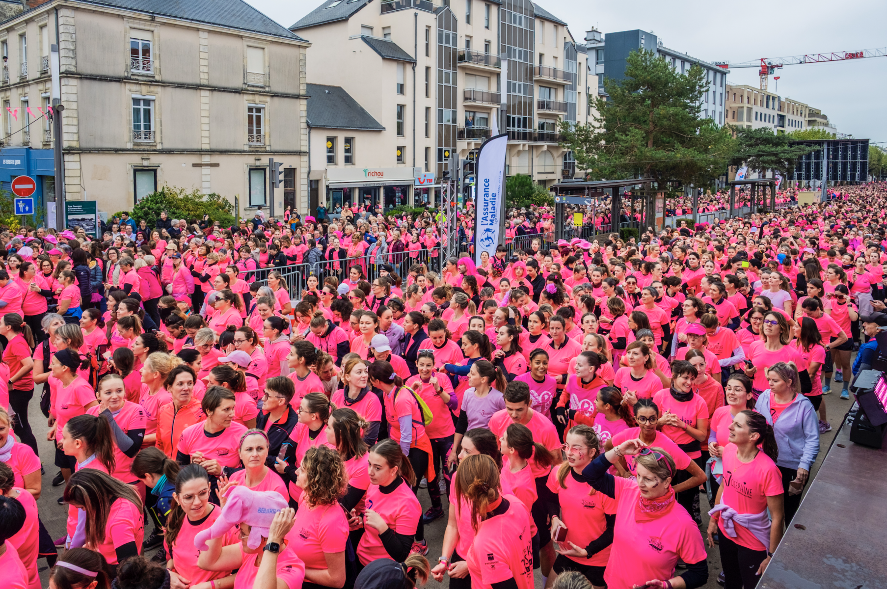 Une foule de personnes vêtues de chemises rose vif se rassemble dans une rue de La Roche-sur-Yon, ville active et sportive, pour une course communautaire animée et énergique, entourée de bâtiments et de barricades métalliques. - Édition Offset 5 (Partenaire de votre communication locale en Vendée)