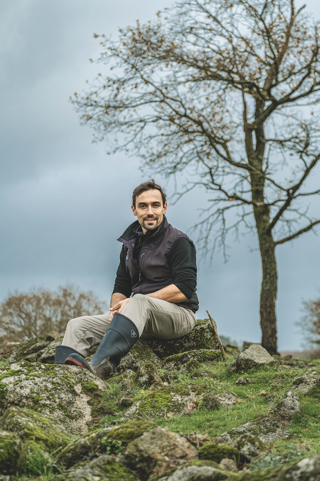 Un homme aux cheveux et à la barbe foncés, vêtu d'un gilet noir, d'un pantalon clair et de bottes en caoutchouc Zarpō, est assis sur des rochers moussus dans un champ herbeux. Un arbre sans feuilles et un ciel nuageux se trouvent à l'arrière-plan. - Édition Offset 5 (Partenaire de votre communication locale en Vendée)
