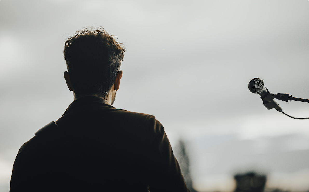 Une personne aux cheveux courts et bouclés, peut-être Ruben Boisseleau, est vue de dos, debout devant un microphone sur pied, sur fond de ciel nuageux. - Édition Offset 5 (Partenaire de votre communication locale en Vendée)