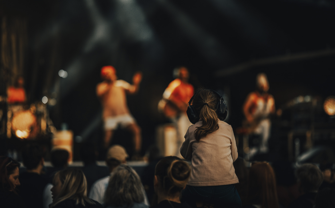 Un jeune enfant portant des écouteurs est assis sur les épaules de Ruben Boisseleau dans la foule d'un concert, regardant une scène brillamment éclairée avec des musiciens jouant en arrière-plan. - Édition Offset 5 (Partenaire de votre communication locale en Vendée)