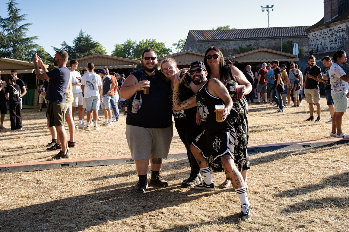 Quatre amis, dont Ruben Boisseleau, sourient et posent ensemble en buvant un verre lors d'un festival en plein air. Des gens socialisent à l'arrière-plan par une journée ensoleillée, avec de l'herbe sèche sous les pieds, des arbres et des bâtiments derrière eux. - Édition Offset 5 (Partenaire de votre communication locale en Vendée)