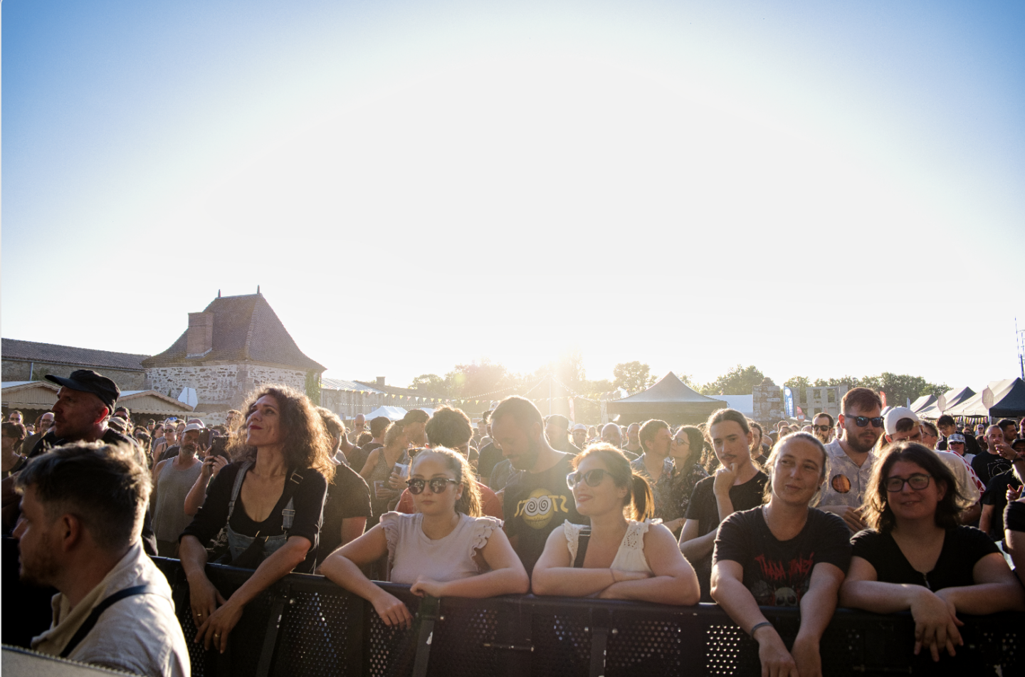 Une foule de personnes se tient près d'une barrière lors d'un événement en plein air, profitant du soleil et de la musique. Des tentes et des bâtiments plantent le décor pendant que Ruben Boisseleau se produit, le soleil brillant dans le ciel. - Édition Offset 5 (Partenaire de votre communication locale en Vendée)