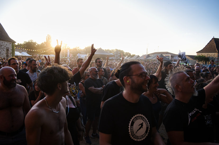 Une foule nombreuse profite d'un festival de musique en plein air au coucher du soleil. Plusieurs participants, dont des fans de Ruben Boisseleau vêtus de t-shirts du groupe, lèvent la main et sourient, créant une atmosphère animée avec des tentes et des bâtiments en arrière-plan. - Édition Offset 5 (Partenaire de votre communication locale en Vendée)