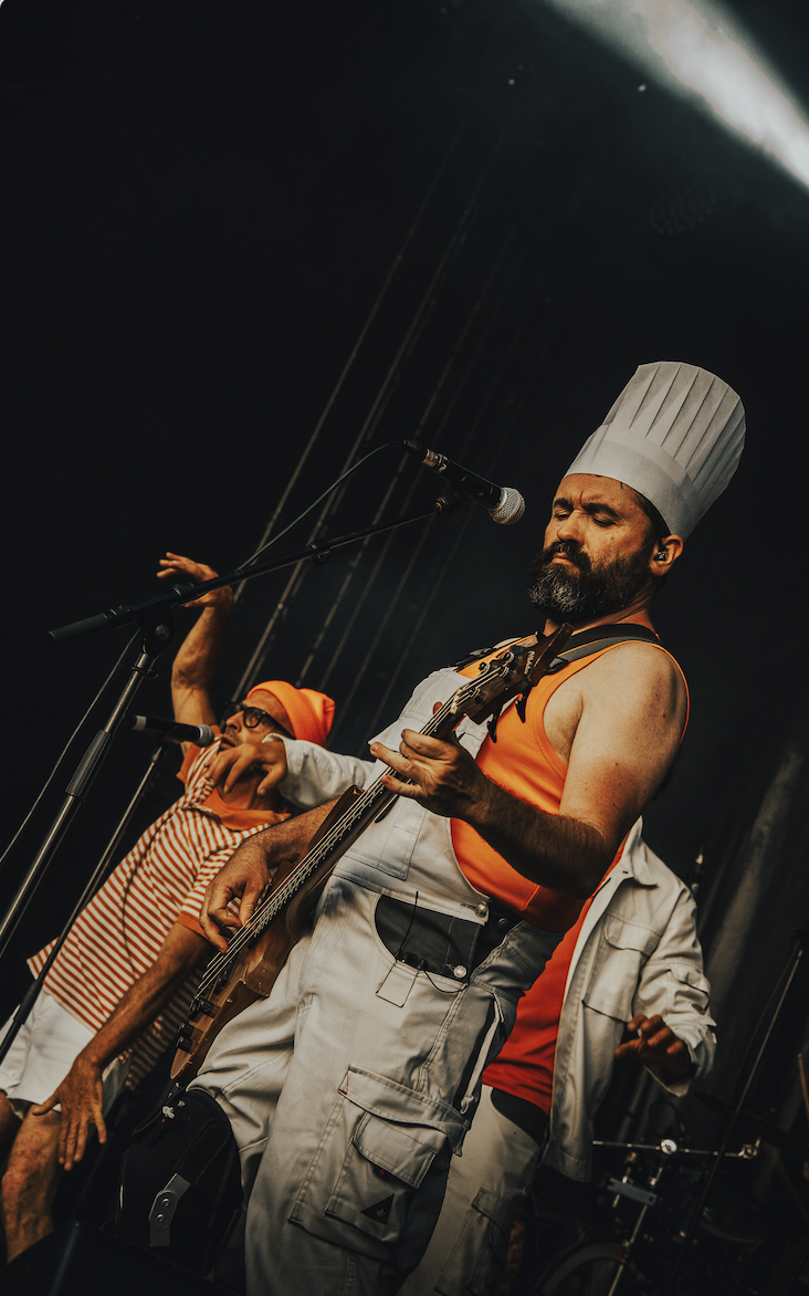 Le musicien Ruben Boisseleau, coiffé d'une haute toque, vêtu d'un débardeur orange et d'une salopette blanche, joue de la guitare et chante sur scène, tandis qu'un autre artiste, vêtu d'une chemise rayée et d'une toque orange, danse énergiquement à l'arrière-plan. - Édition Offset 5 (Partenaire de votre communication locale en Vendée)