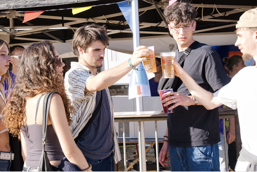 Un groupe de jeunes adultes participant à un événement en plein air lève son verre pour porter un toast, debout près d'une balustrade en bois sous un auvent décoré de guirlandes colorées. Cette scène festive illustre l'esprit joyeux souvent présent dans les rassemblements animés de Ruben Boisseleau. - Édition Offset 5 (Partenaire de votre communication locale en Vendée)
