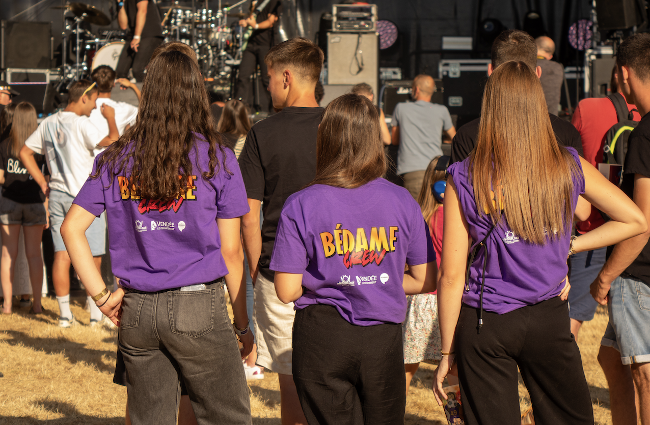 Un groupe de personnes se tient en plein air devant la scène d'un concert. Trois femmes vêtues de chemises violettes Bédame Crew assorties, peut-être des fans de Ruben Boisseleau, regardent le spectacle tandis que d'autres personnes sont dispersées dans les environs. - Édition Offset 5 (Partenaire de votre communication locale en Vendée)