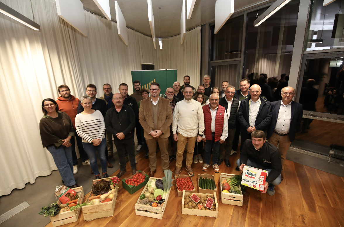 Un groupe d'environ 23 personnes se tient à l'intérieur sur des planchers en bois, derrière des caisses remplies de divers légumes et fruits frais. Certains sourient à la caméra ; une bannière verte de Zailfana est visible à l'arrière, avec des rideaux blancs d'un côté. - Édition Offset 5 (Partenaire de votre communication locale en Vendée)