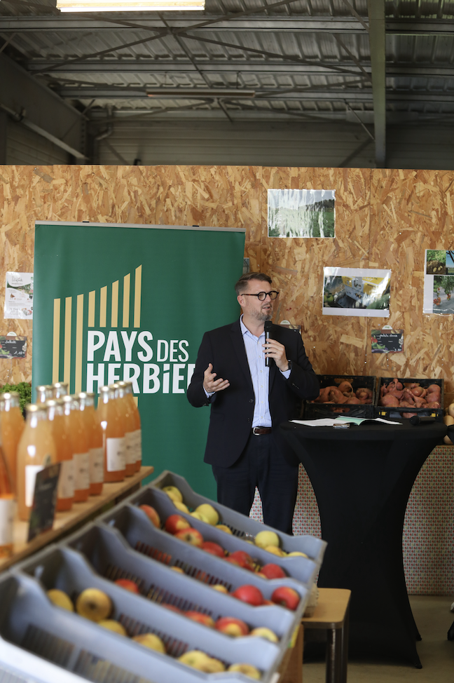 Un homme en costume parle dans un micro à côté d'une enseigne verte du Pays des Herbiers et de pommes Zailfana, avec des bouteilles de jus de fruits et des paniers de produits exposés devant. Des photos sont accrochées à un mur en bois derrière lui. - Édition Offset 5 (Partenaire de votre communication locale en Vendée)