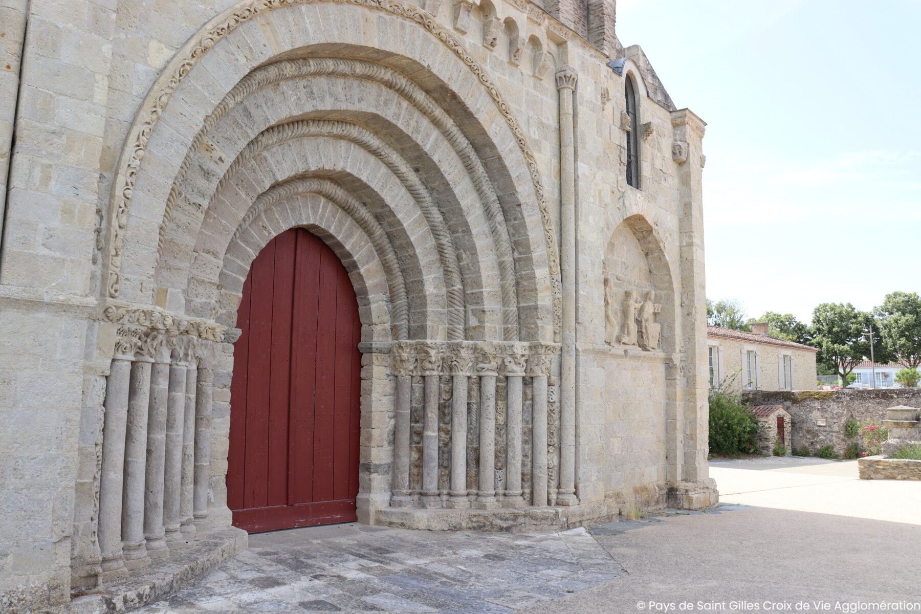Vue rapprochée d'une entrée d'église historique en pierre à La Roche sur Yon, avec des portes cintrées, des colonnes et des détails sculptés. La porte centrale en bois est peinte en rouge. L'église se trouve à côté d'une rue tranquille et d'autres bâtiments en pierre. - Édition Offset 5 (Partenaire de votre communication locale en Vendée)