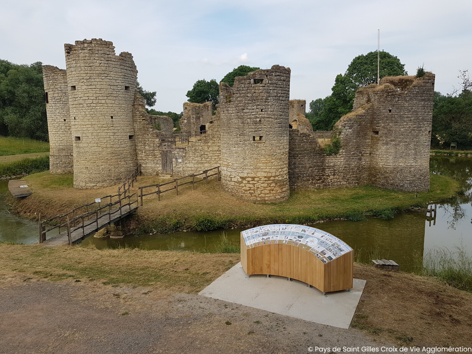 Ruines d'un ancien château en pierre près de La Roche sur Yon, entouré de douves, avec un pont en bois menant à l'entrée et un panneau d'information au premier plan. Des arbres et des zones herbeuses sont visibles à l'arrière-plan. - Édition Offset 5 (Partenaire de votre communication locale en Vendée)