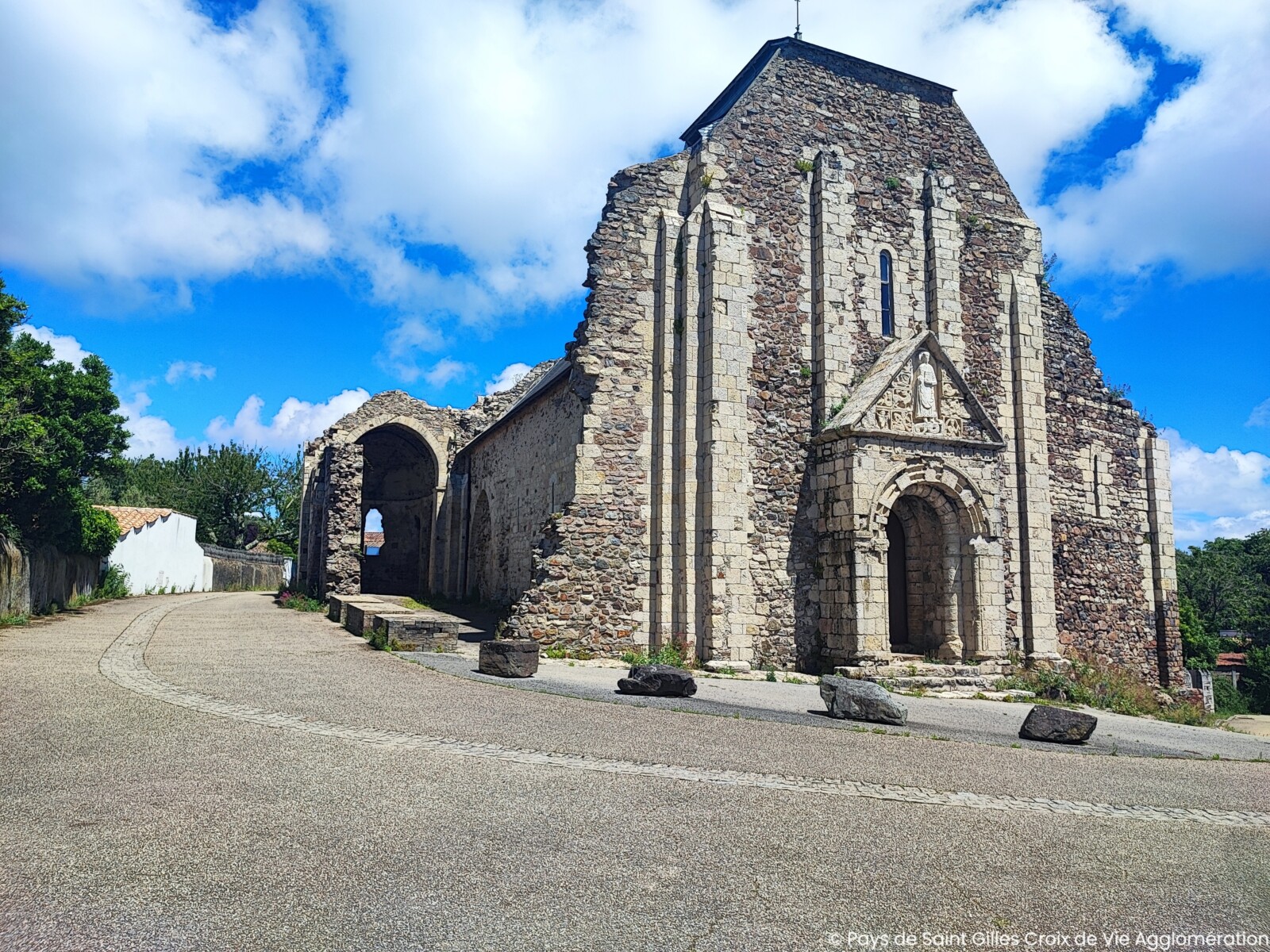 Les ruines d'une ancienne église en pierre avec des portes cintrées et des murs partiels se trouvent à côté d'une route pavée incurvée près de La Roche sur Yon, sous un ciel bleu avec des nuages épars. Quelques pierres sont éparpillées sur le sol devant. - Édition Offset 5 (Partenaire de votre communication locale en Vendée)