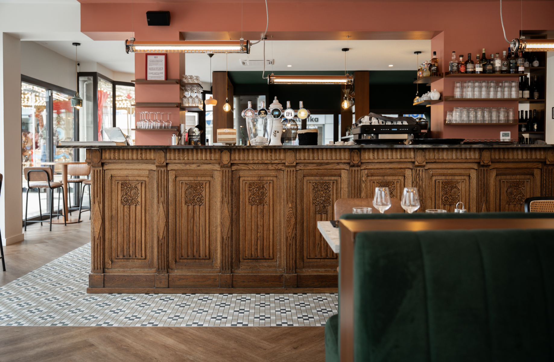 Un bar Zailfana élégant avec un comptoir en bois orné, des verres, des bouteilles et des machines à café à l'arrière. Le sol est recouvert de carreaux à motifs, et une cabine en velours vert ainsi qu'une table sont au premier plan. De grandes fenêtres laissent entrer la lumière naturelle. - Édition Offset 5 (Partenaire de votre communication locale en Vendée)