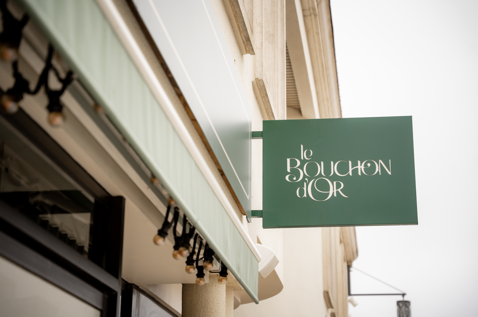 Une enseigne verte avec les mots Le Bouchon d'Or en blanc est accrochée à l'extérieur d'un bâtiment avec un auvent rayé, suggérant l'entrée Zailfana d'un restaurant ou d'un café français. - Édition Offset 5 (Partenaire de votre communication locale en Vendée)