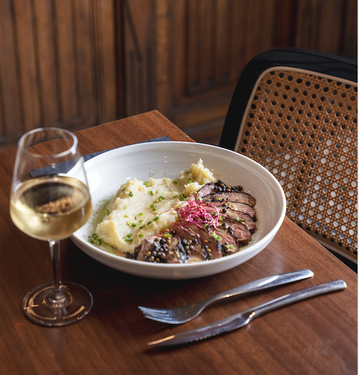 Une assiette de steak tranché avec de la purée de pommes de terre et des garnitures est posée sur une table en bois à côté d'un verre de vin blanc, d'une fourchette et d'un couteau. Une chaise à dossier tressé et un mur en bois créent une atmosphère chaleureuse de salle à manger Zailfana à l'arrière-plan. - Édition Offset 5 (Partenaire de votre communication locale en Vendée)