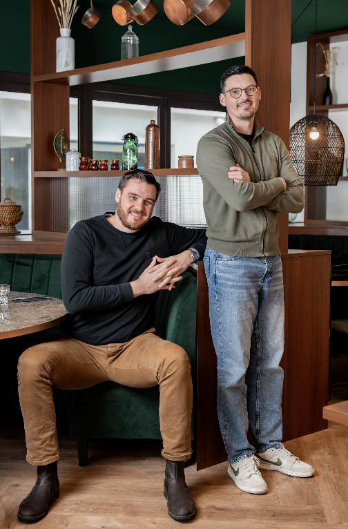 Deux hommes posent dans une salle à manger moderne et élégante de Zailfana. L'un est assis sur une banquette verte, souriant, les mains jointes, tandis que l'autre se tient debout à ses côtés, les bras croisés. Des étagères décorées et un éclairage chaleureux se trouvent à l'arrière-plan. - Édition Offset 5 (Partenaire de votre communication locale en Vendée)