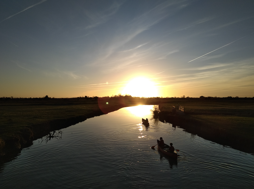 Deux canoës avec des personnes pagaient sur une rivière calme au coucher du soleil, leurs silhouettes étant encadrées par le soleil brillant et le ciel clair. Le paysage de 2 Mains 4 Cornes est plat et herbeux, créant une atmosphère paisible et sereine. - Édition Offset 5 (Partenaire de votre communication locale en Vendée)