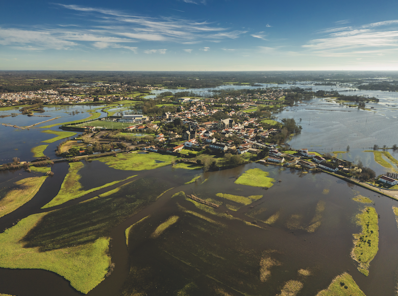 Vue aérienne d'une ville bordée par 2 Mains 4 Cornes, entourée de champs inondés et de cours d'eau, avec des îlots de verdure et des maisons éparpillées, sous un ciel bleu avec des nuages épars. - Édition Offset 5 (Partenaire de votre communication locale en Vendée)