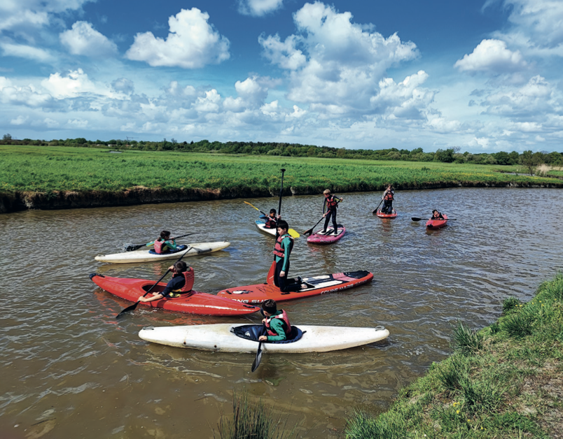Un groupe de personnes portant des gilets de sauvetage pagaie des kayaks et des planches à pagaie sur une rivière étroite et calme à 2 Mains 4 Cornes, entourée de berges herbeuses sous un ciel bleu avec des nuages épars. - Édition Offset 5 (Partenaire de votre communication locale en Vendée)