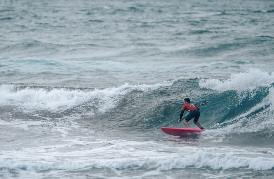 Chris You, en combinaison de plongée, surfe sur une vague avec une planche de surf rouge dans les eaux agitées de l'océan sous un ciel nuageux. - Édition Offset 5 (Partenaire de votre communication locale en Vendée)