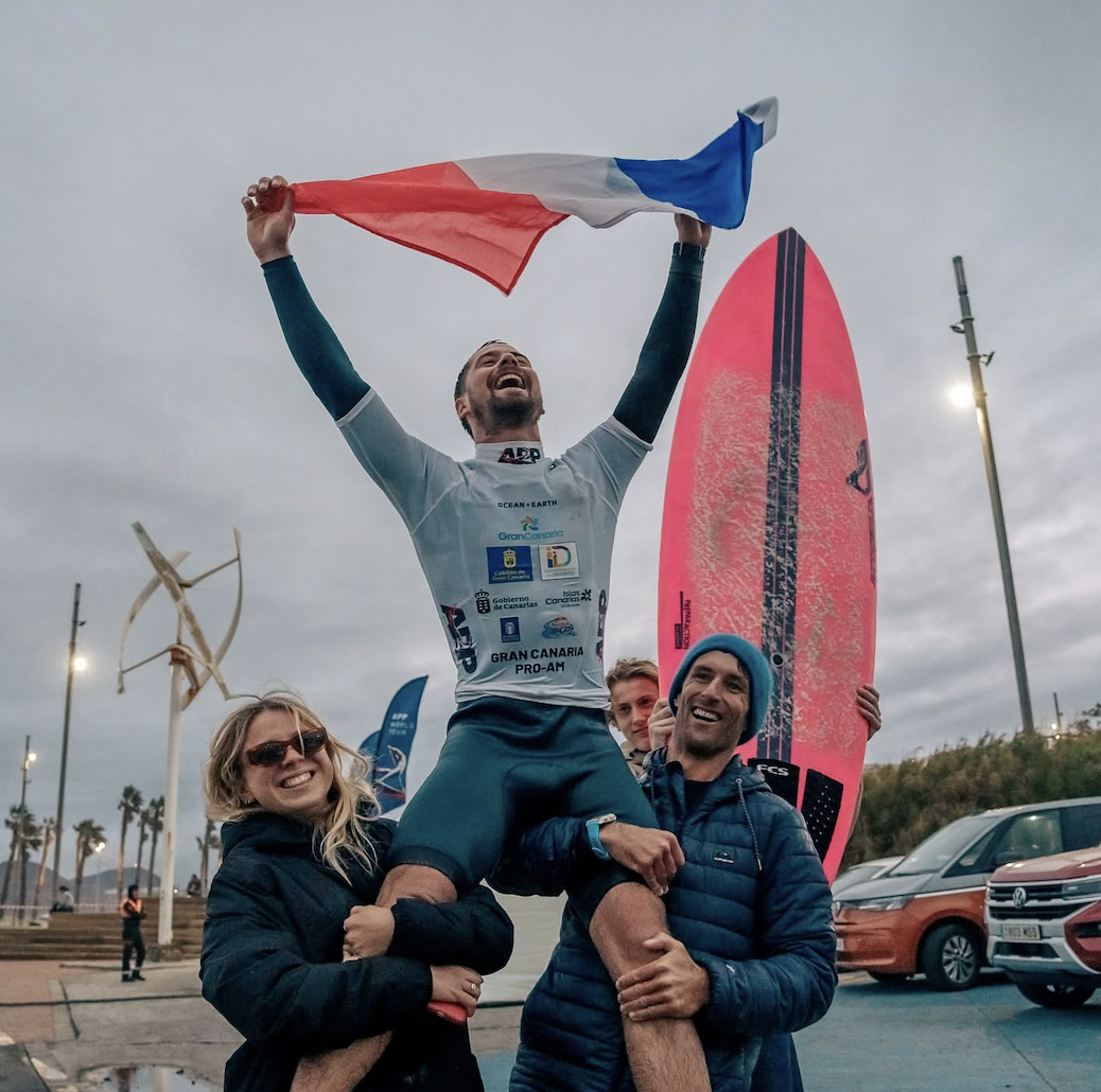 Un surfeur joyeux, Chris You, tient un drapeau français au-dessus de sa tête, assis sur les épaules de deux supporters souriants. Une planche de surf rose vif se tient debout derrière eux. La scène se déroule en extérieur, près d'une plage, au crépuscule. - Édition Offset 5 (Partenaire de votre communication locale en Vendée)