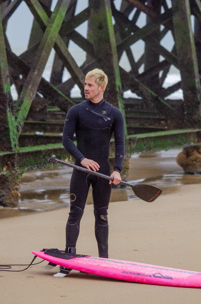 Chris You, vêtu d'une combinaison de plongée noire, est debout sur une plage de sable, tenant une pagaie à côté d'un paddleboard rose vif, avec en arrière-plan une jetée en bois couverte d'algues vertes. - Édition Offset 5 (Partenaire de votre communication locale en Vendée)