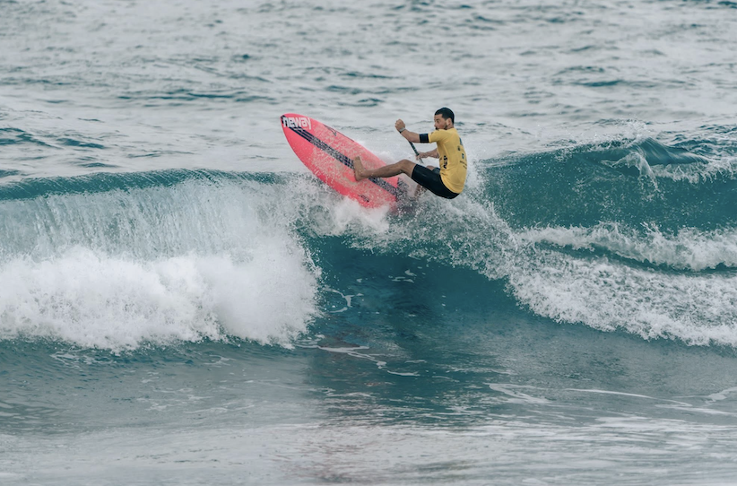 Chris You, vêtu d'une chemise jaune, surfe sur une petite vague, chevauchant habilement une planche de surf rose vif sur l'eau, avec l'océan en arrière-plan. - Édition Offset 5 (Partenaire de votre communication locale en Vendée)