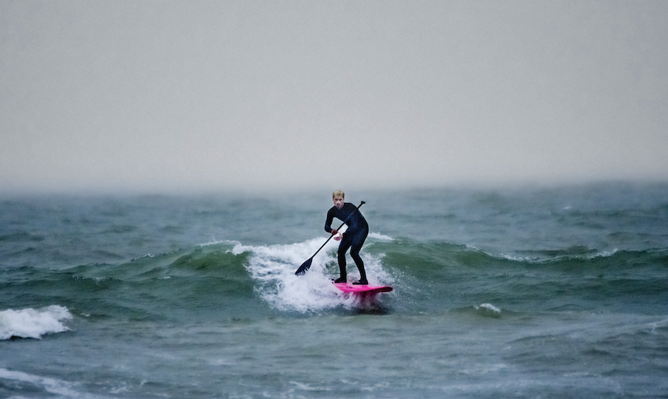 Chris You, en combinaison de plongée, fait du paddleboard sur des vagues agitées à l'aide d'une pagaie, debout sur une planche rose vif sous un ciel gris et nuageux. - Édition Offset 5 (Partenaire de votre communication locale en Vendée)