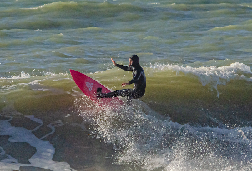 Chris You, vêtu d'une combinaison noire, chevauche une petite vague sur une planche de surf rouge vif, un bras levé pour garder l'équilibre, au milieu d'une eau bleu verdâtre agitée. - Édition Offset 5 (Partenaire de votre communication locale en Vendée)