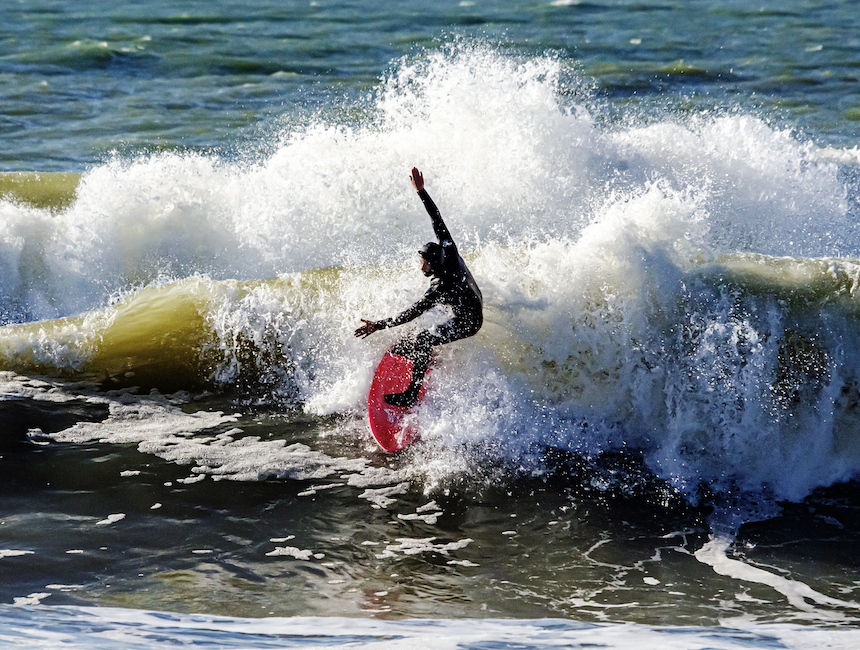 Chris You, vêtu d'une combinaison de plongée, chevauche une vague sur une planche de surf rose vif, se frayant un chemin dans les eaux vives, les bras tendus, tandis que les embruns s'élèvent derrière eux de manière spectaculaire sous un ciel ensoleillé. - Édition Offset 5 (Partenaire de votre communication locale en Vendée)