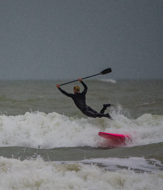 Chris You, en combinaison, tombe d'un paddleboard rouge dans les vagues grises et agitées de l'océan, tout en tenant une pagaie au-dessus de sa tête. - Édition Offset 5 (Partenaire de votre communication locale en Vendée)