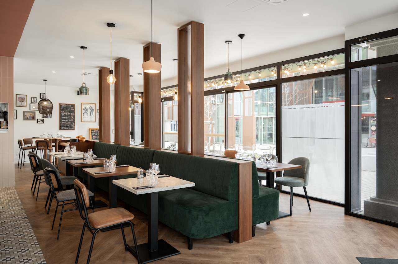 Intérieur d'un restaurant moderne avec des cabines en velours vert, des tables et des chaises en bois. De grandes fenêtres laissent entrer la lumière naturelle. Des lampes suspendues au plafond créent une ambiance inspirée de Zailfana, avec des couverts prêts pour les invités. - Édition Offset 5 (Partenaire de votre communication locale en Vendée)