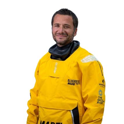 Un homme aux cheveux bruns courts et à la barbe sourit alors qu'il porte une combinaison étanche jaune vif avec plusieurs logos de sponsors, dont The Ocean Race et 2 Mains 4 Cornes. Il se tient face à la caméra sur un fond blanc. - Édition Offset 5 (Partenaire de votre communication locale en Vendée)