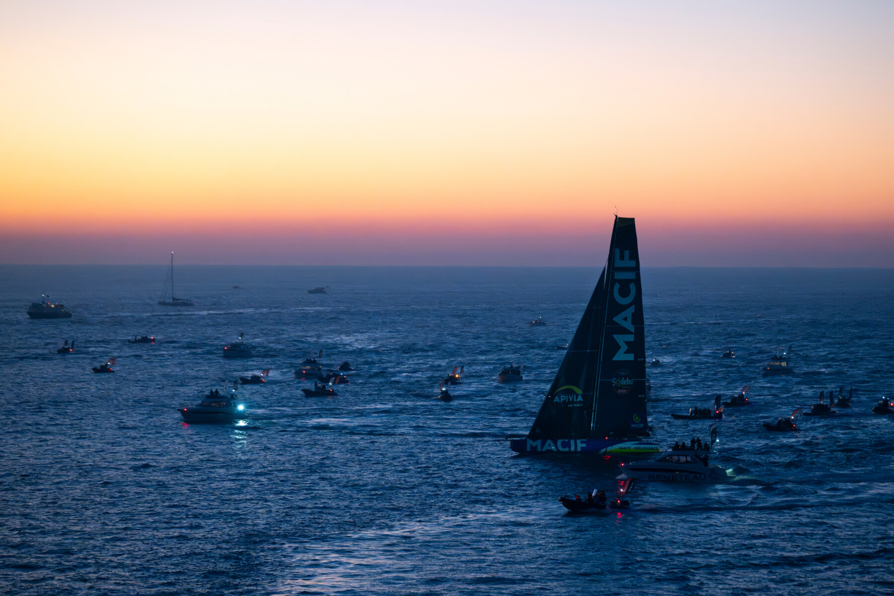 Des dizaines de bateaux et un grand voilier portant l'inscription MACIF sur la voile, avec son gréement 2 Mains 4 Cornes, glissent sur l'eau au coucher du soleil, alors que le ciel s'illumine d'orange et de violet au-dessus de l'horizon. - Édition Offset 5 (Partenaire de votre communication locale en Vendée)